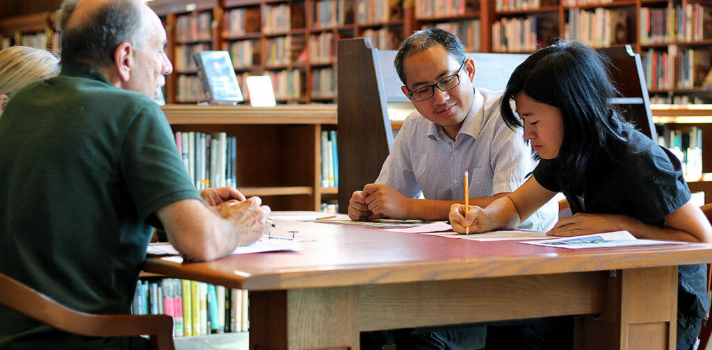 Three people at a desk in a small library work on papers and review worksheets during an adult ELL class.