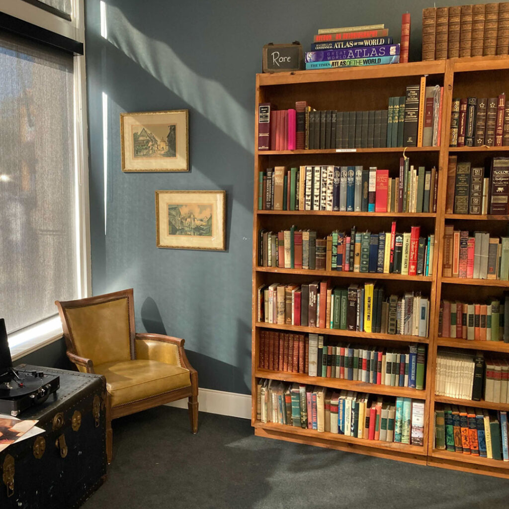 A large bookshelf sits near a comfy chair against a blue wall with gray carpet on a sunny day outside the window.