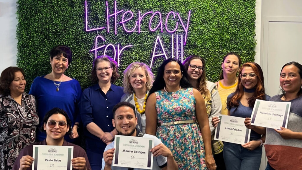 Photo of 9 English Language Learning students with 1 instructor and 2 volunteers celebrating their 2022-2023 year. They are holding certificates in front of a "Literacy for All" sign.