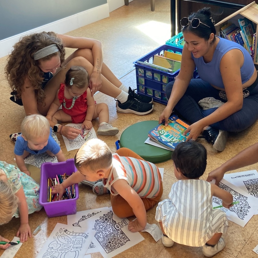 A group of kids play on the floor as two adult volunteers watch